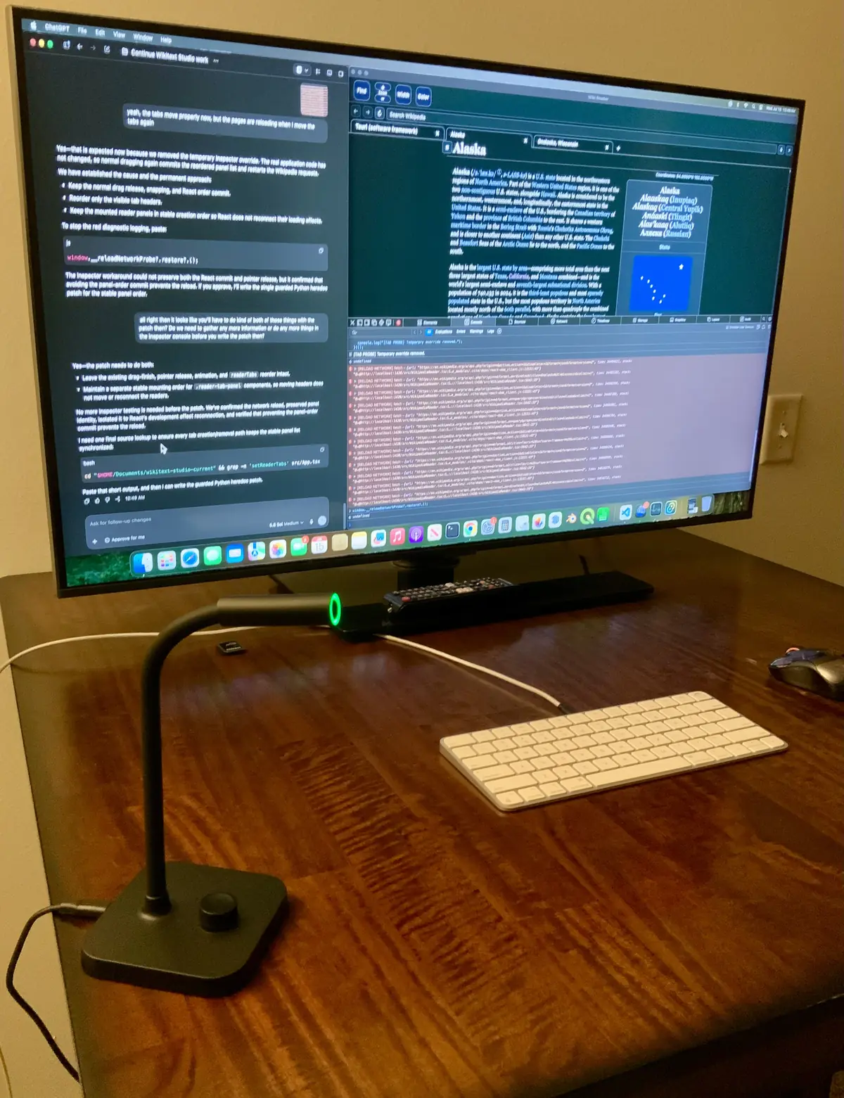 Mikrofon meja berleher lentur dengan lampu hijau di depan monitor besar yang menampilkan percakapan dengan agen coding dan panel inspeksi, keyboard putih di atas meja kayu